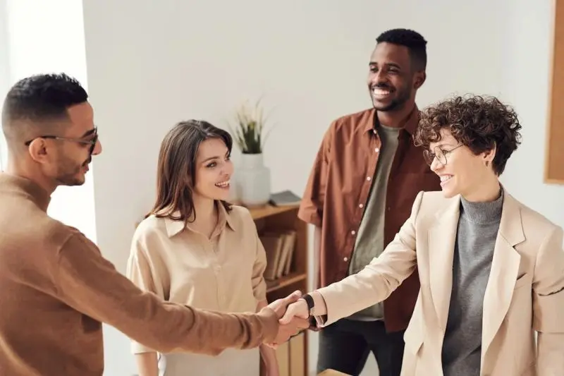 Quatre personnes souriantes dans un environnement professionnel, deux d’entre elles se serrant la main en signe de collaboration.