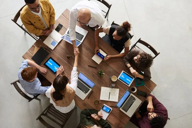 Vue aérienne d’un groupe de personnes travaillant ensemble autour d’une table en bois, avec des ordinateurs portables, des tablettes, des carnets et des téléphones. Deux personnes se serrent la main au centre de la table.