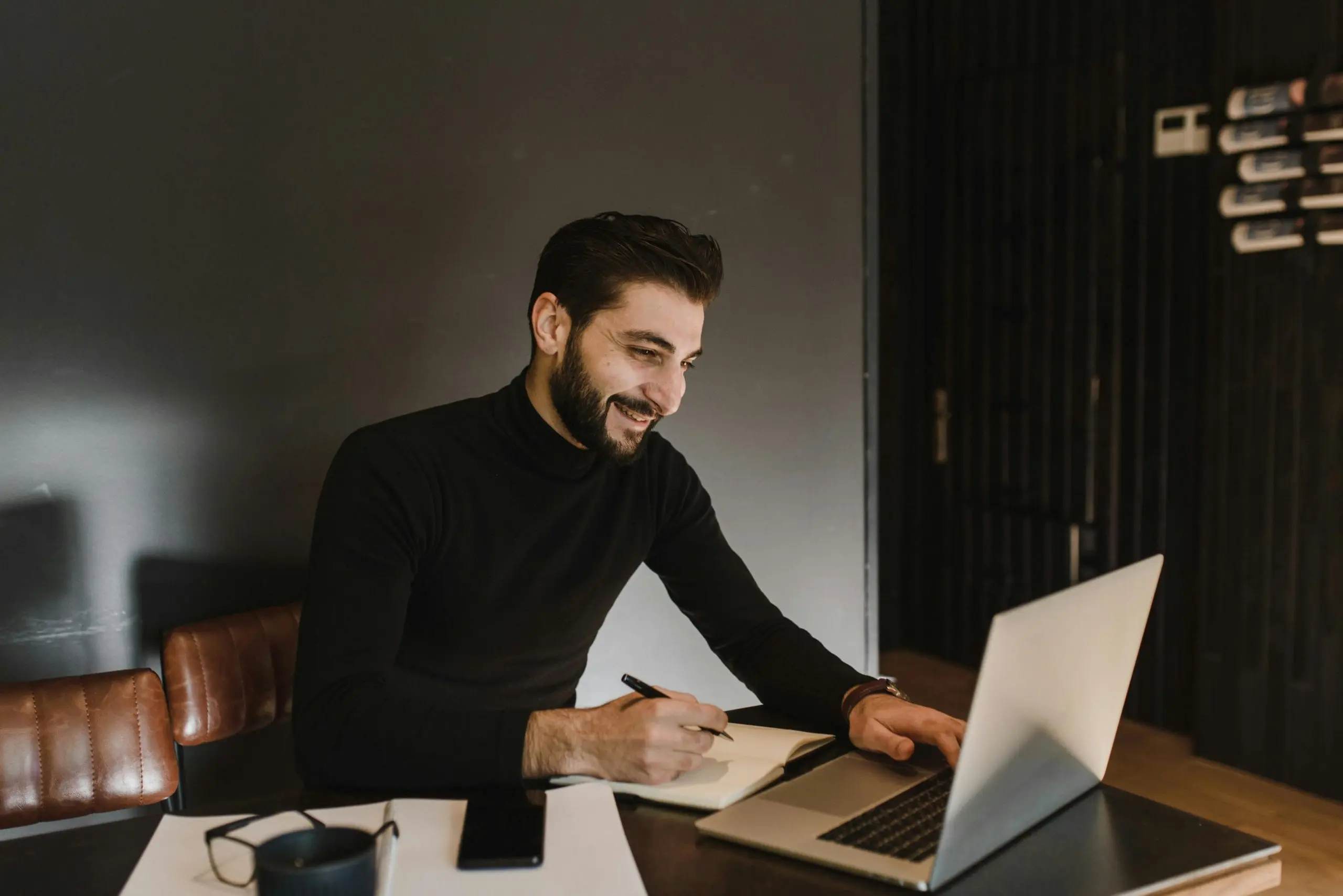 Un homme souriant, vêtu d’un pull noir, assis à une table, prenant des notes dans un carnet tout en travaillant sur un ordinateur portable.