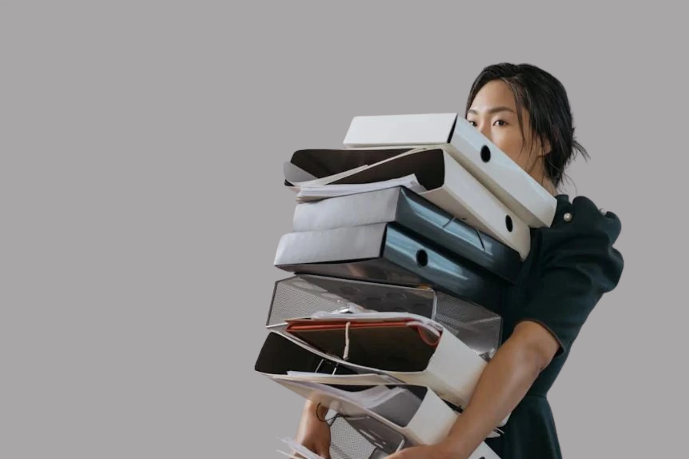 Une personne portant une robe noire tient une pile de dossiers et de classeurs contre un fond gris.